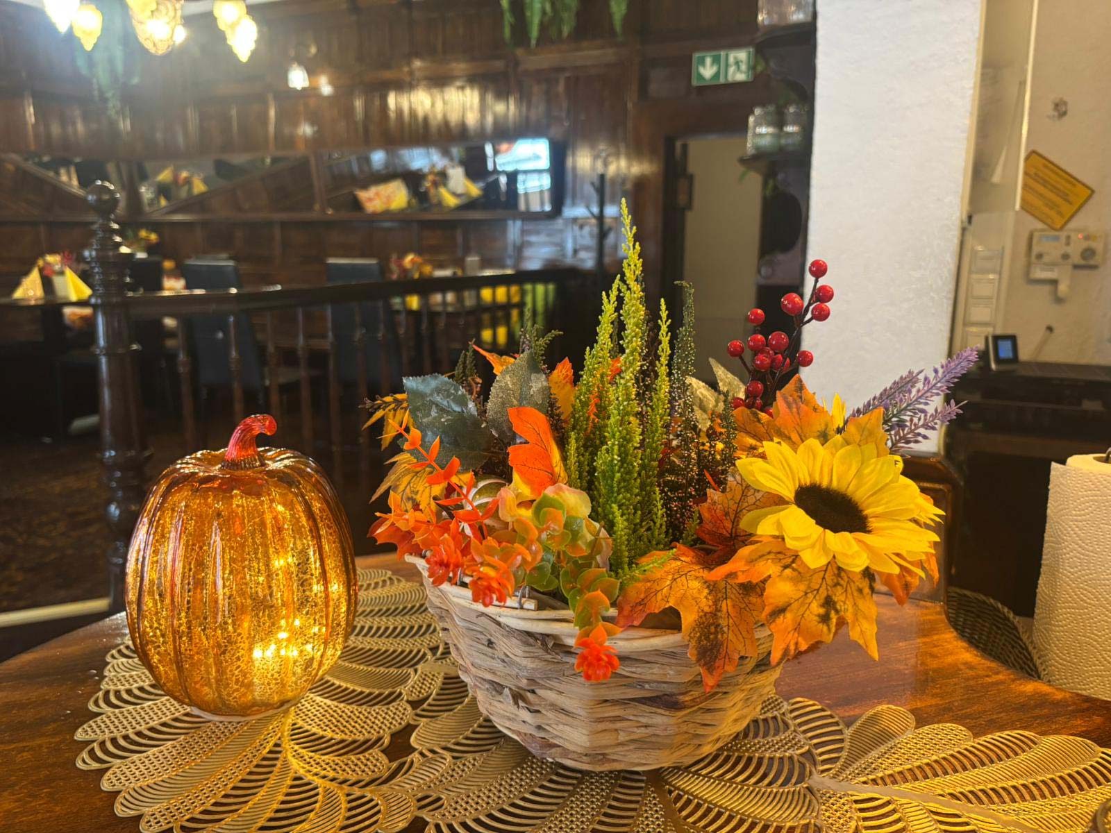 Ein herbstliches Gesteck mit Sonnenblume und bunten Blättern steht neben einer leuchtenden Kürbis Laterne auf einem Holztisch